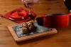 A hand is drizzling chocolate sauce over two cannoli on a slate serving board, with a red pot and lid in the background. Produktbild-Vorschau