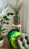 Several round cushions in various shades of green are stacked in the foreground, with a potted plant and a vase with dried flowers in the background. Produktbild-Vorschau