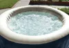 An inflatable hot tub filled with bubbling water, set outdoors on a paved patio with grass and plants in the background. Produktbild-Vorschau