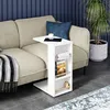 A white side table with shelves is placed next to a sofa in a living room setting. A roll of paper, a book, and a small figurine are on the shelves. A tablet and a cup of coffee are on the top surface of the table. Anteprima dell’immagine prodotto