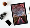 A framed picture of a canal lined with cherry blossoms at night, placed on a white surface next to a vintage camera, a smartphone, a cup of tea, and a pencil. Produktbild-Vorschau