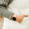 A close-up shot of a woman wearing a smartwatch on her wrist, with her hand tucked into the pocket of a white puffer jacket. She is also wearing a grey knitted sweater. Produktbild-Vorschau