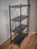 A black metal shelving unit with four shelves is shown in a room with wooden flooring and a light-colored wall. Produktbild-Vorschau