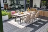 An outdoor dining set with chairs and a table on a deck, next to a grill and greenery. Produktbild-Vorschau