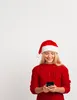 A young woman wearing a Santa hat and a red sweater is looking at her phone. Podgląd zdjęcia produktu