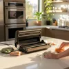 A vacuum sealer is shown on a kitchen counter with food items in bags, and a person's hands are visible arranging the food. Anteprima dell’immagine prodotto