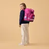A young girl wearing a backpack stands against a plain background. Produktbild-Vorschau