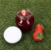 A jar of strawberry jam with a strawberry slice on top, a white lid, and two whole strawberries on a grassy surface. Produktbild-Vorschau