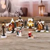 A collection of illuminated Christmas village houses and figures arranged on a wooden surface, with artificial snow scattered around. Produktbild-Vorschau