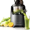 A Kuvings slow juicer is shown in operation, with green juice being dispensed into a glass. Fresh fruits and vegetables are arranged around the juicer. Podgląd zdjęcia produktu
