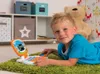 A young boy is lying on a green rug, playing with a toy laptop. He is looking at the camera and smiling. In the background, there is a shelf with storage boxes and a large teddy bear. Produktbild-Vorschau