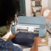 A person is using a Cricut Maker 4 cutting machine to cut a dark blue material. Produktbild-Vorschau