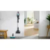 A cordless vacuum cleaner is mounted on a white wall next to a wooden bench. In the foreground, a floor cleaning attachment and two cleaning pads are visible. Náhled obrázku produktu