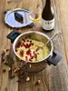 A cooking pot filled with couscous, berries, and nuts, placed on a wooden table next to a bottle of wine and a glass of white wine. Produktbild-Vorschau