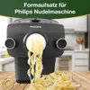 A close-up view of a Philips pasta maker attachment extruding tagliatelle pasta onto a wooden table. A pile of freshly made pasta is visible in the foreground. The background shows a blurred kitchen scene. Voir les images du produit