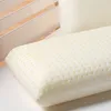 A close-up shot of a white latex pillow with many small holes, resting on a white surface. In the background, wooden slats of a bed frame are visible. Produktbild-Vorschau