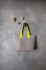 A grey woven tote bag with bright yellow handles hangs from a dark red hook on a textured grey wall. Several other decorative hooks in various colors are also visible on the wall. Produktbild-Vorschau