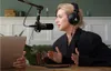 A woman wearing headphones is speaking into a microphone, with a laptop in front of her. Produktbild-Vorschau