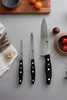 Three kitchen knives are laid out on a gray surface, with some food items and kitchenware in the background. Náhled obrázku produktu