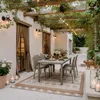 An outdoor patio dining set with chairs and a table, covered by a pergola with hanging lights and vines. A cat is walking on the patio. Produktbild-Vorschau