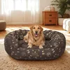 A golden retriever dog is lying comfortably in a round, brown dog bed with a paw print pattern. The bed is placed on a beige carpet in a well-lit room with a window and wooden furniture in the background. Podgląd zdjęcia produktu