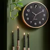 A black cuckoo clock with a wooden frame hangs on a green wall, next to a hanging plant and four black candles in candle holders. Produktbild-Vorschau
