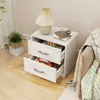 A white nightstand with two drawers is placed next to a sofa with a blanket. A lamp and a mug are on top of the nightstand. A plant is on the left side of the nightstand. Podgląd zdjęcia produktu