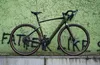 A dark green gravel e-bike is parked in front of a green wall with graffiti. Produktbild-Vorschau
