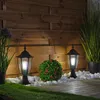 Two vintage-style outdoor lanterns are illuminated in a garden setting at dusk, surrounded by white stones, green grass, a decorative bush with red flowers, and tall green plants. Náhľad obrázku produktu