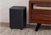 A black subwoofer sits on a rug next to a wooden cabinet. Produktbild-Vorschau