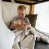 A young child sits under a table, playing with a stuffed animal and a blanket. Produktbild-Vorschau