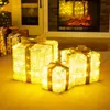 Three illuminated gift boxes with golden bows are placed on a floor in front of a fireplace and a doorway. Podgląd zdjęcia produktu