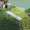 A white folding bench is set up on a grassy area next to a body of water, with camping chairs and a cooler nearby. Produktbild-Vorschau