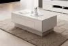 A modern white coffee table with a drawer, placed on a beige rug in a living room setting. Podgląd zdjęcia produktu