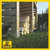 A close-up view of a wooden log store with firewood stacked inside, set in a grassy outdoor environment. Produktbild-Vorschau