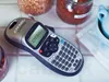 A DYMO LetraTag label maker is shown on a tiled surface, with jars of spices and a blue bowl in the background. Produktbild-Vorschau