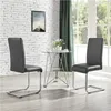 Two dark gray faux leather chairs are placed around a small glass-top dining table with a metal frame. The background features a white brick wall with a framed picture and a dark green curtain. A bright window is visible in the background. Anteprima dell’immagine prodotto