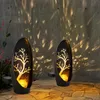 Two solar-powered garden lights with a tree design are placed on a stone patio in front of a textured wall. Produktbild-Vorschau
