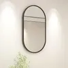 A modern oval mirror with a black frame hangs on a white wall, with soft lighting from above. A small plant is visible in the lower left corner. Náhled obrázku produktu