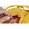A close-up of a hand interacting with a strap and a ring on a yellow backpack. Produktbild-Vorschau