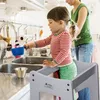 A young child stands on a learning tower in a kitchen, playing with water from the faucet. Produktbild-Vorschau