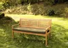 A wooden garden bench with a green cushion is placed on a grassy lawn in a park-like setting. Produktbild-Vorschau