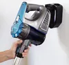 A hand holds a cordless stick vacuum cleaner against a white wall. The vacuum cleaner is blue and gray with a black battery pack attached to the wall mount. Produktbild-Vorschau