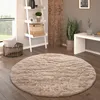 A round, shaggy rug is placed on a wooden floor in a living room setting. A ladder shelf with decorative items, a basket with a pillow, and a plant are visible in the background. Anteprima dell’immagine prodotto