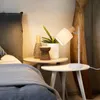 A bedside table with a lamp, vase, and plant next to a bed. Produktbild-Vorschau