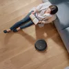 A person is reading a book on the floor next to a sofa, while a robot vacuum cleaner operates nearby. Náhled obrázku produktu