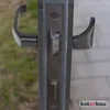 A close-up view of a door handle mechanism and escutcheon plates, with a blurred background of grass and paving stones. Náhľad obrázku produktu