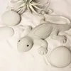 A light gray concrete lizard figurine is placed on a white sandy surface, surrounded by smooth, light gray stones and a small air plant. Produktbild-Vorschau