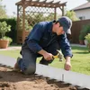 A man is kneeling in a garden, using a hammer to install white garden edging into the soil. The edging is flexible and has metal stakes attached. Produktbild-Vorschau