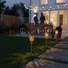A backyard patio at dusk with solar-powered flame lights illuminating a pathway, a grill smoking in the background, and two men standing near a dining table. Náhled obrázku produktu